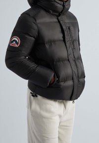 Veste matelassée noire avec un col haut, dotée d'une poche zippée sur le côté gauche de la poitrine et d'un patch logo sur la manche. Design texturé et matelassé.