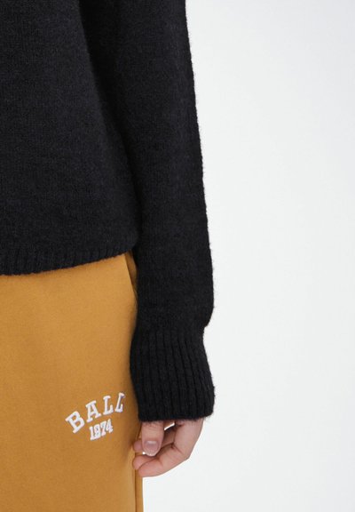 Zwarte trui met een ribgebreide zoom en manchetten, gemaakt van een zachte, gestructureerde stof. Gecombineerd met mosterdkleurige broek met witte geborduurde tekst.