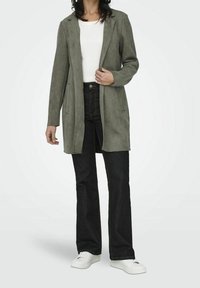 Olivgrüner Wildlederblazer mit tailliertem Schnitt, Revers mit Kerbe, Fronttaschen und langen Ärmeln, getragen über einem weißen Hemd und schwarzen Jeans.