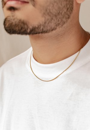 Goldene Kettenhalskette auf einem nackten Hals, die über einem weißen Baumwoll-T-Shirt ruht. Die Kette ist dünn und glatt, mit einem subtilen Glanz.