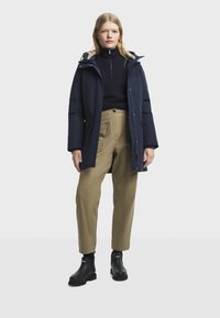 Žena u tamnoplavom zimskoj kaputu s kapuljačom, crnom puloveru s četvrtim patent-zatvaračem, bež hlačama i crnim gležnjačama, stoji uz bijelu pozadinu.