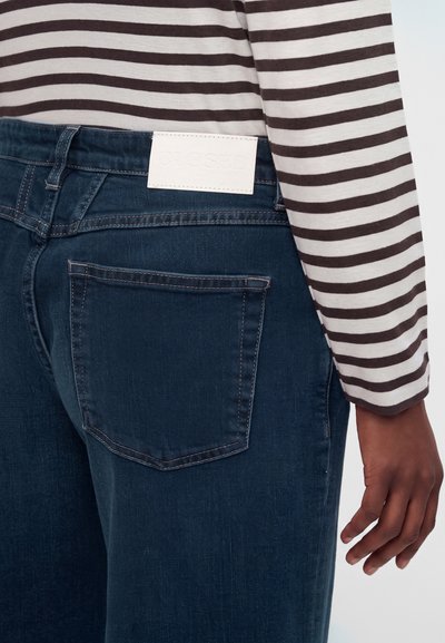 Personne portant un jean bleu foncé avec une étiquette blanche sur la ceinture et une chemise à manches longues rayée marron et blanche.