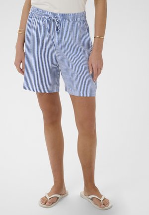 Vrouw die blauwe en witte verticale gestreepte korte broek met trekkoord draagt, een wit topje, gouden armbanden en witte slippers, staand voor een effen achtergrond.