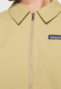 Patagonia zip-up jacket in light beige, featuring a front zipper, large collar, and embroidered logo. The fabric has a smooth texture.