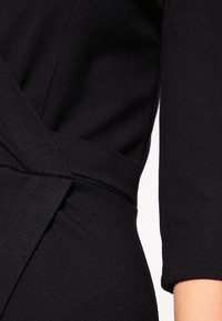 Black wrap dress featuring a fitted bodice, three-quarter sleeves, and a textured fabric. The waist is accentuated with a fabric tie.