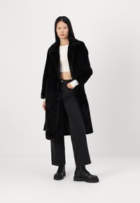 Abrigo de piel sintética negra con cuello con muescas, mangas largas y bolsillos laterales. Se lleva sobre un top blanco corto y se combina con pantalones negros de pierna ancha.