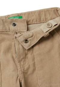 Béžové kordové kalhoty s knoflíkem a patentkou, nastavitelným pasem s knoflíkem a částečně viditelnou zelenou značkovou etiketou.