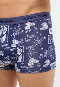 Gros plan sur un boxer Björn Borg bleu marine avec des illustrations blanches à thème tropical et océanique sur la hanche et la cuisse d'une personne.