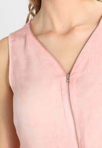 Haut sans manches rose avec un décolleté en V et un détail de fermeture éclair métallique. Le tissu semble léger avec une texture lisse. Aucun motif ni accent observé.