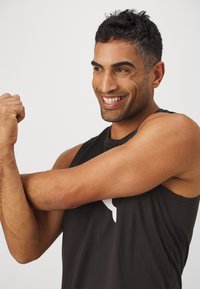 Zwarte mouwloze sportshirt met een ronde hals, gladde stof en een wit logo aan de voorkant. Het model strekt zijn arm uit.