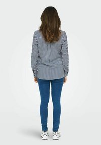 Blusa a righe in blu navy e bianco, maniche lunghe, polsini con bottoni e orlo arrotondato, abbinata a jeans blu aderenti e sneakers bianche.