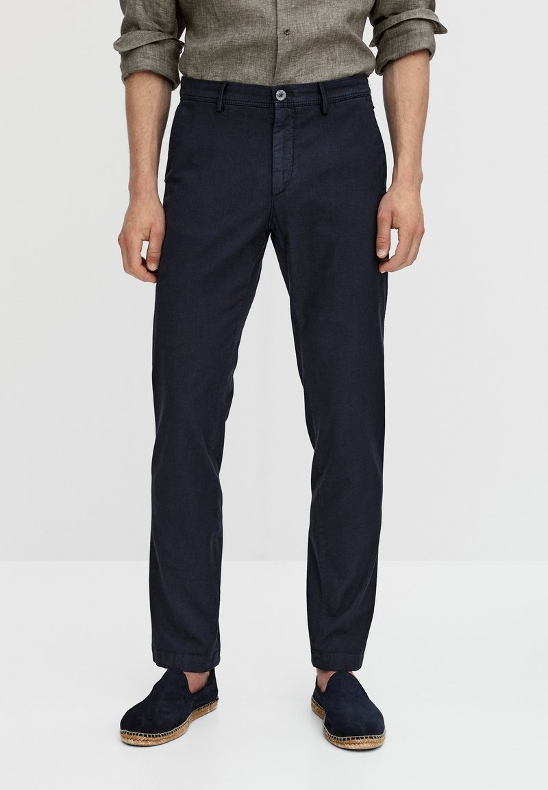 Tummansininen kevyt housut, joissa on suora leikkaus, sileä pinta, nappikiinnitys ja etutaskut; paritettu tummien liukuvien kenkien kanssa.