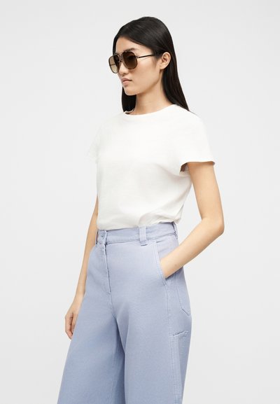 Γυναίκα που φορά καφέ γυαλιά ηλίου, λευκό μπλουζάκι και ανοιχτό μπλε παντελόνι με φαρδιά μπατζάκια και ψηλή μέση, με το ένα χέρι στην τσέπη, σε απλό φόντο.