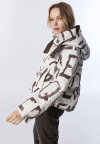 Femme blonde en veste à capuche blanche et marron à motifs et pantalon sombre pose de profil sur fond clair.