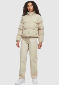 Veste matelassée beige courte avec col montant, fermeture éclair frontale et poignets élastiques, assortie à un pantalon droit beige clair et des baskets blanches.