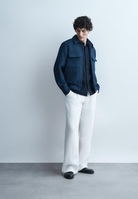 Muž s kučeravými vlasmi v tmavomodrom saká, čiernom tričku, bielych širokých nohaviciach a čiernych topánkach stojí pri svetlosivej stene.