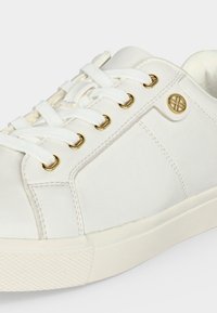 Baskets basses blanches avec lacets blancs, œillets dorés et un logo circulaire doré sur le côté au-dessus de la semelle.
