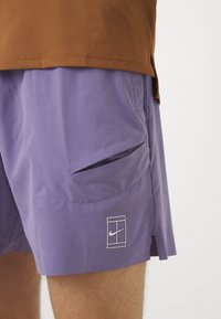 Lilla idrettsshorts med sidelomme, en liten hvit Nike-logo, og et lett, pustende stoff. Brun mesh-topp synlig ovenfor.
