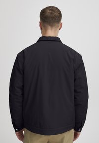 Svart jakke med et enkelt design, med krage og knappede mansjetter, laget av glatt stoff, sett fra baksiden.