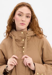 Brown hooded jacket with gold hardware, featuring a soft lining, drawstring neck, and snap closure. Model holds jacket at the collar.