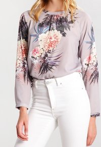 Blomstret bluse i lysegrå med livlige pink og lilla blomster, lange ærmer, opsamlet halsudskæring, stylet med hvide højtaljede jeans.