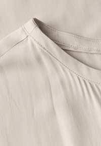 Tessuto beige con una trama liscia, dotato di uno scollo rotondo e di una leggera arricciatura nella cucitura della spalla. I bordi cuciti sono visibili.