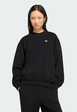 Ung kvinde med langt mørkt hår iført en sort Adidas sweatshirt med logo, hænder i lommerne, neutralt udtryk, lysegrå baggrund.