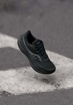 Zwarte Saucony hardloopschoen met reflecterend logo en Gore-Tex label die zweeft boven een gestructureerde asfalt ondergrond met witte verfmarkeringen.