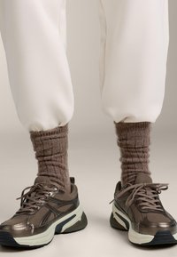 Sneakers a rete marroni con accenti chiari, abbinate a calzini marroni a coste e morbidi. Pantaloni jogger color crema, raccolti alle caviglie.