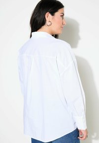 Camicia bianca a maniche lunghe realizzata in tessuto leggero, con colletto classico e orlo curvo. Indossata in modo casual con jeans.