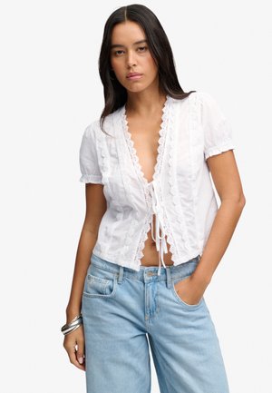 Mujer con cabello largo y oscuro lleva una blusa blanca de manga corta con detalle de encaje y lazos en el frente, combinada con vaqueros azul claro y pulseras plateadas.