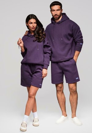 Lila Baumwollhoodies und Shorts mit Kordelzügen und Logoakzenten, getragen von zwei Personen vor einem neutralen Hintergrund.