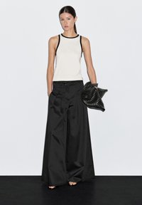 Black wide-leg pants paired with a cream tank top featuring black trim. Model holds a textured black clutch. Plain background.