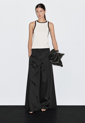 Black wide-leg pants paired with a cream tank top featuring black trim. Model holds a textured black clutch. Plain background.
