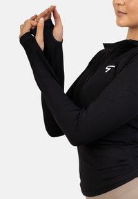 Crna majica dugih rukava s ovratnikom na munje, koja ima otvore za palce na manžetama i uski je kroj, izrađena od glatkog, rastezljivog materijala.