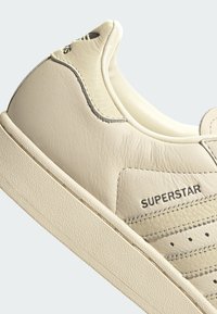 Αθλητικό παπούτσι Adidas Superstar σε εκρού δέρμα με πλευρική λεπτομέρεια με διάτρητες ρίγες και κείμενο "SUPERSTAR" στο επάνω μέρος.