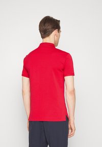 Červené polo tričko s žebrovaným límcem, krátkými rukávy a rozparky po stranách u lemu pro snadnější pohyb. Vyrobeno z hladké bavlněné tkaniny.