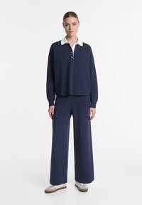 Navy Strickpullover mit weißem Kragen und Knopfverschluss, kombiniert mit lockeren, navyblauen Hosen und hellen Sneakers.