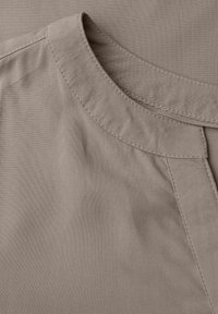 Beige kangas, jossa on sileä rakenne, pyöreä pääntie ja ommeltu yksityiskohta. Kankaassa on hienovarainen kiilto, ja se laskeutuu pehmeästi.