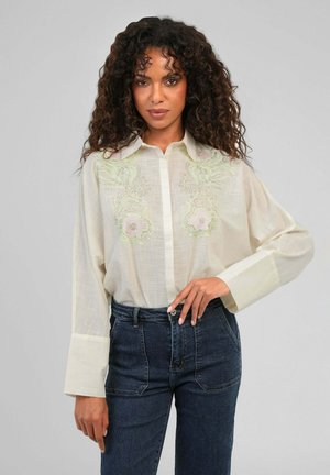 Femme aux cheveux bouclés portant un chemisier écru transparent avec broderie florale et un jean taille haute foncé, posant devant un fond uni.