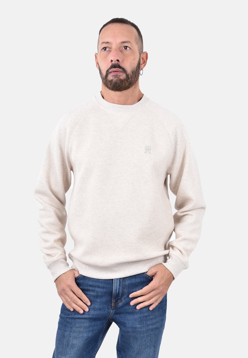 Felpa beige chiaro con maniche raglan, polsini e orlo a costine, con un piccolo logo sul petto. Indossata con jeans blu.