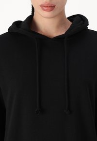 Μαύρο hoodie με κουκούλα και κορδόνι, μαλακή υφή και άνετη εφαρμογή. Το ύφασμα φαίνεται λείο με ομοιόχρωμη απόχρωση.