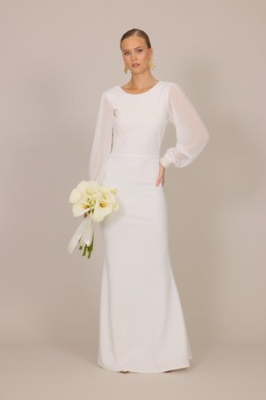 Femme en robe blanche à manches longues tenant un bouquet de callas blancs, portant des boucles d'oreilles dorées imposantes, debout devant un fond beige.