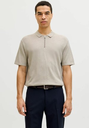 Mann iført beige poloshorte med korte ermer og frontglidelås, stukket inn i mørkeblå bukser med svart belte, stående mot en ensfarget bakgrunn.