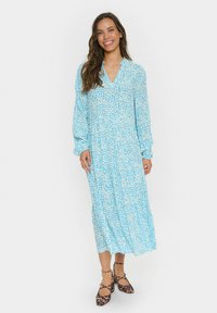 Lång blå klänning med vitt abstrakt mönster, V-ringning och långa ärmar, matchad med leopardmönstrade skor. Lätt, flödande material.
