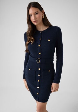 Donna che indossa un vestito in maglia blu navy aderente con bottoni dorati, cintura e tasche frontali, in piedi davanti a uno sfondo grigio uniforme.
