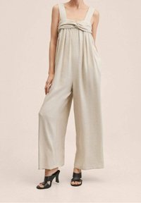 Combinaison beige avec jambes larges, décolleté carré et détail noué à l'avant. Fabriquée en tissu doux, elle dispose de deux poches latérales. Chaussures noires à talons hauts.