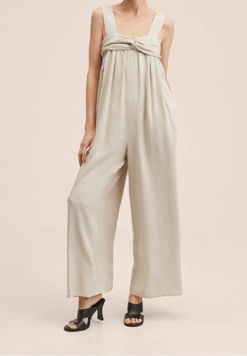 Combinaison beige avec jambes larges, décolleté carré et détail noué à l'avant. Fabriquée en tissu doux, elle dispose de deux poches latérales. Chaussures noires à talons hauts.