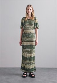 Abito lungo con strisce ondulate verdi, beige e crema. Maniche a sbuffo e silhouette aderente. Indossato con sandali neri, caratterizzati da una suola spessa.