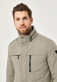 Light gray padded jacket with a high collar, two zippered chest pockets, and black accents on shoulders. Smooth texture with a sleek design.